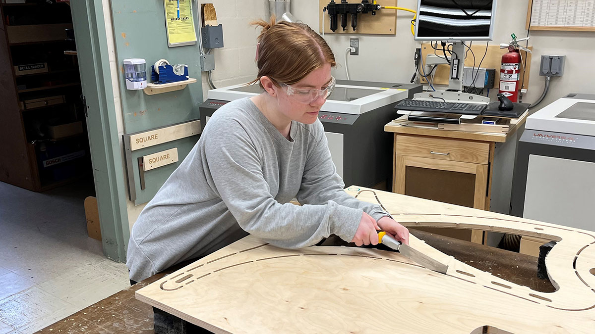 In a workshop with machinery and a computer, a young woman wearing safety glasses and a gray sweatshirt traces a metal tool over a wooden board with cut-out designs.
