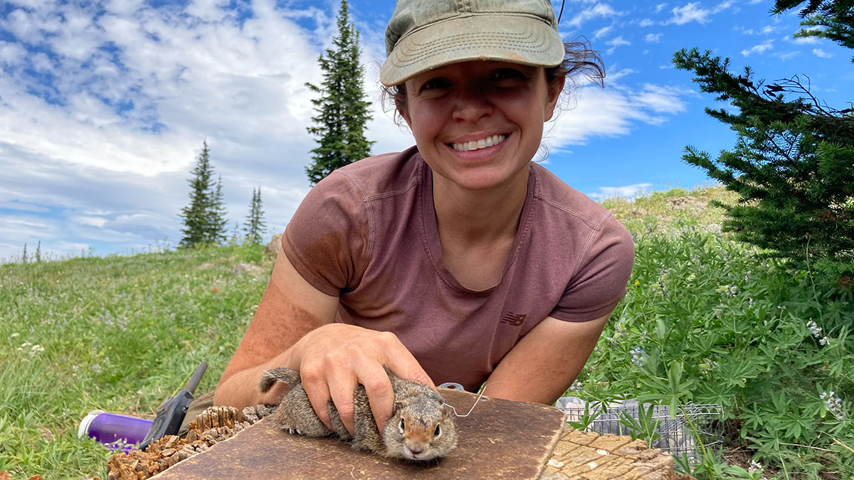 A young woman smiles as she holds a Northern Idaho ground squirrel in an open field dotted with pine trees under a blue sky.
