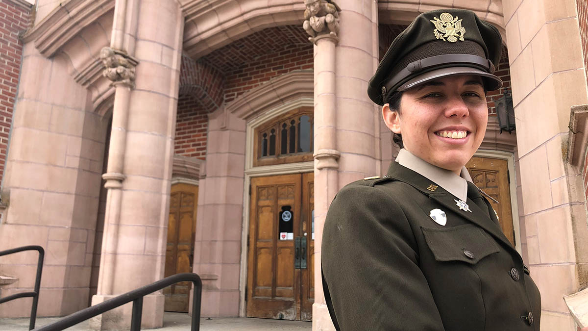 Woman in Army green dress uniform stands in front of brick building.