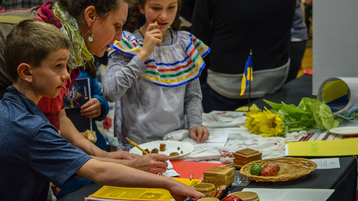 A small crowd and children inspect decorated Easter eggs (Pysanky), faux sunflowers, Ukrainian nesting dolls, as well as several carved wooden boxes.  