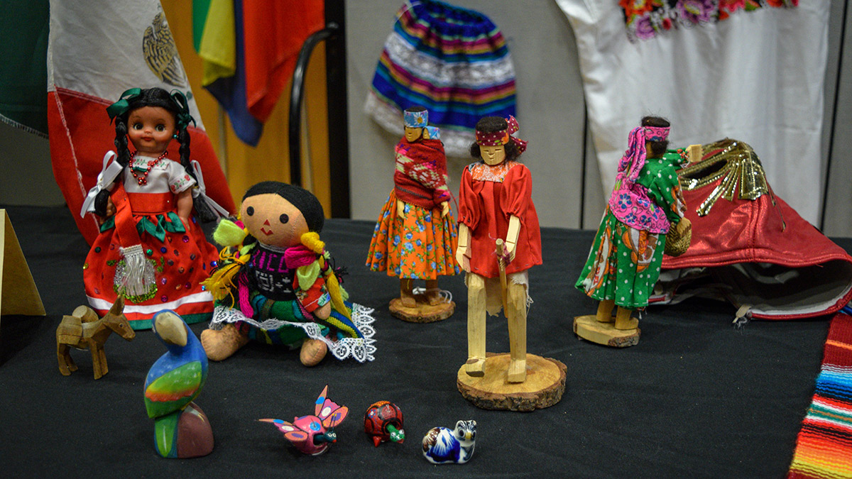 Five dolls wearing various traditional Mexican clothing. There is also a stand with the Mexican flag, along with carved and painted animal figurines including a donkey, a parrot, a butterfly and a turtle.