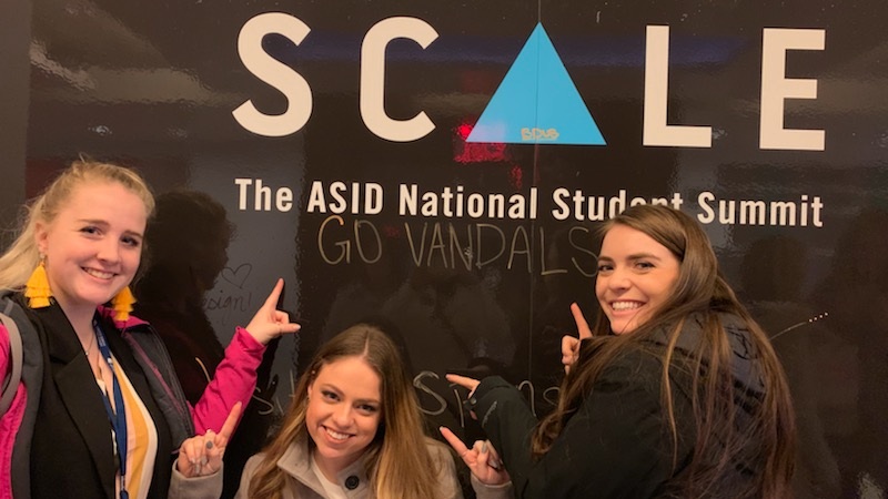 Three students pose in front of an event poster at the ASID National Student Summit.