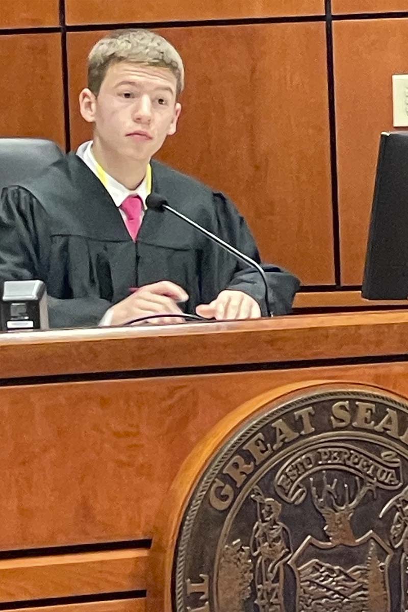 A young man wearing a judges robe sits behind a podium in a courtroom.