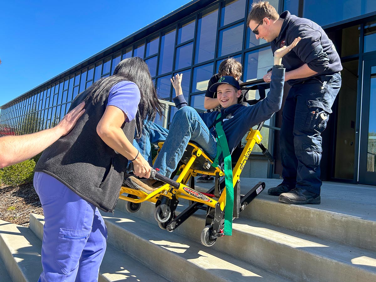Young boy gets carried down the stairs in a chair by EMTs.