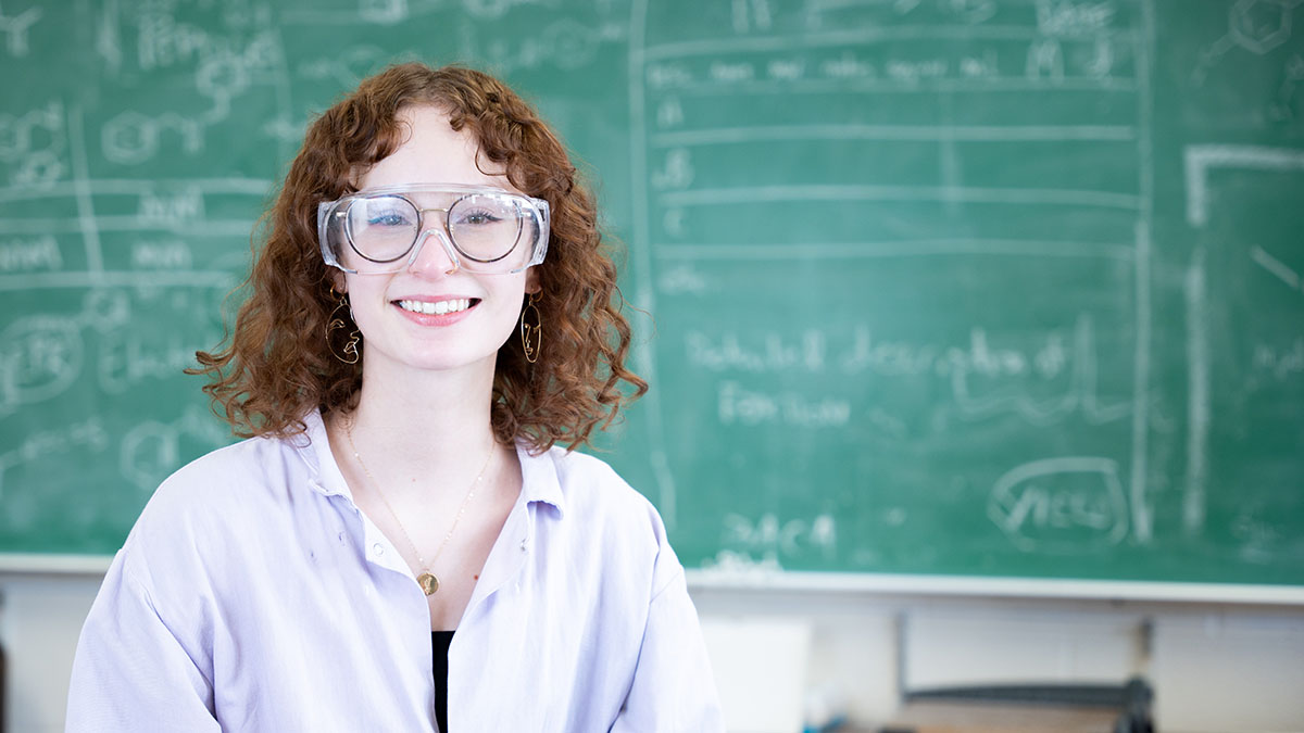 Woman with curly hair wears safety glasses and a lab coat.