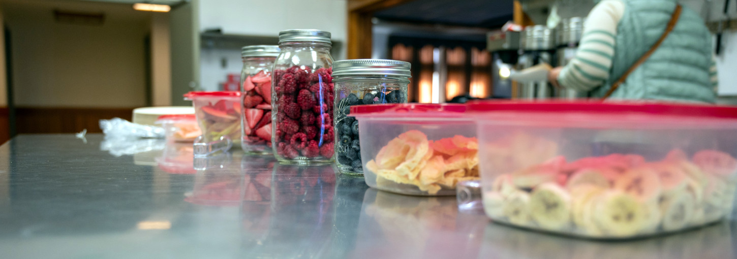 Containers of dehydrated bananas, mangoes, blueberries, raspberries and other fruit on a counter. 