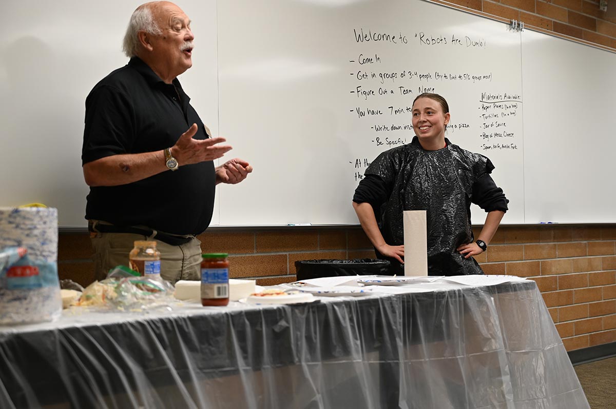 A U of I student wearing a trash bag smiles as a professor speaks in front of a table of supplies to make a pizza in a coding activity.