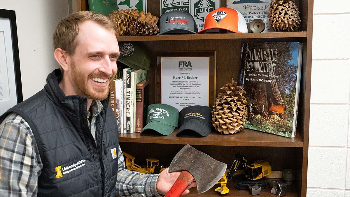 Smiling man holds axe in front of bookshelf of logging memorabilia.