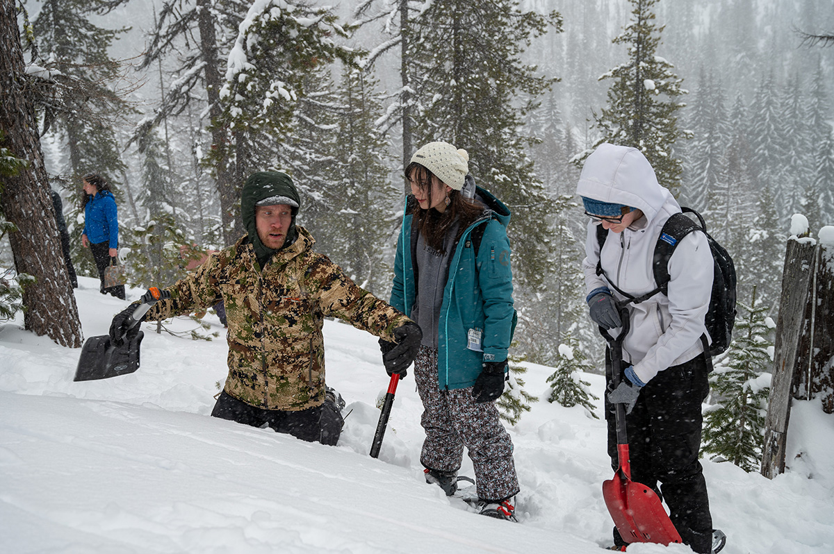 Man indicates with his arms while two students look on in snow field.