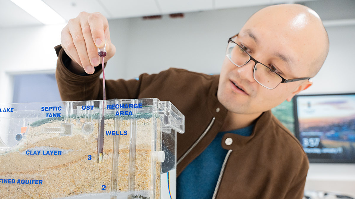 Meng Zhao, a bald man wearing glasses and a brown jacket over a blue T-shirt, inserts a small rod into a plastic tank with different layers of labeled soil. 