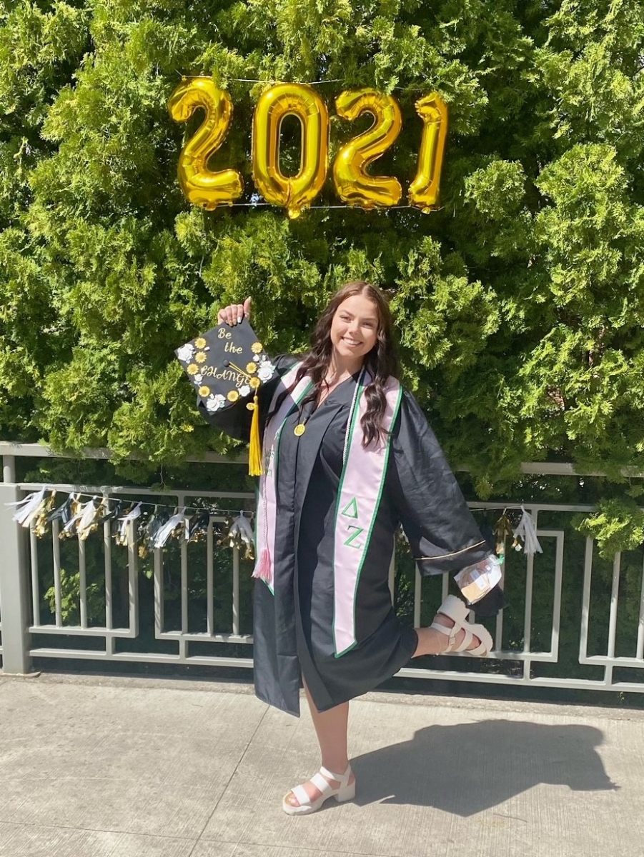 Emma Arman stands in front of trees featuring balloons spelling “2021”. She is wearing a graduation gown and holding up her cap, which reads “Be the Change.”