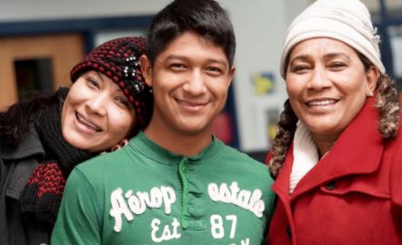 A teen boy wearing a green shirt stands between a girl near his age leaning her head towards his and an adult woman wearing a red coat and white hat.