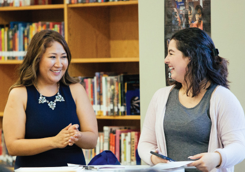 Two women smile at each other. One wears a dressy tank top and the other wears a cardigan. They have papers and pens spread out in front of them and library books in the background.