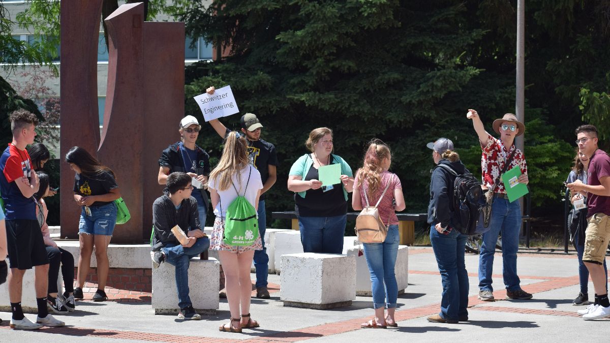 A dozen teens gather on a plaza around a young man holding up a "Schweitzer Engineering" sign.