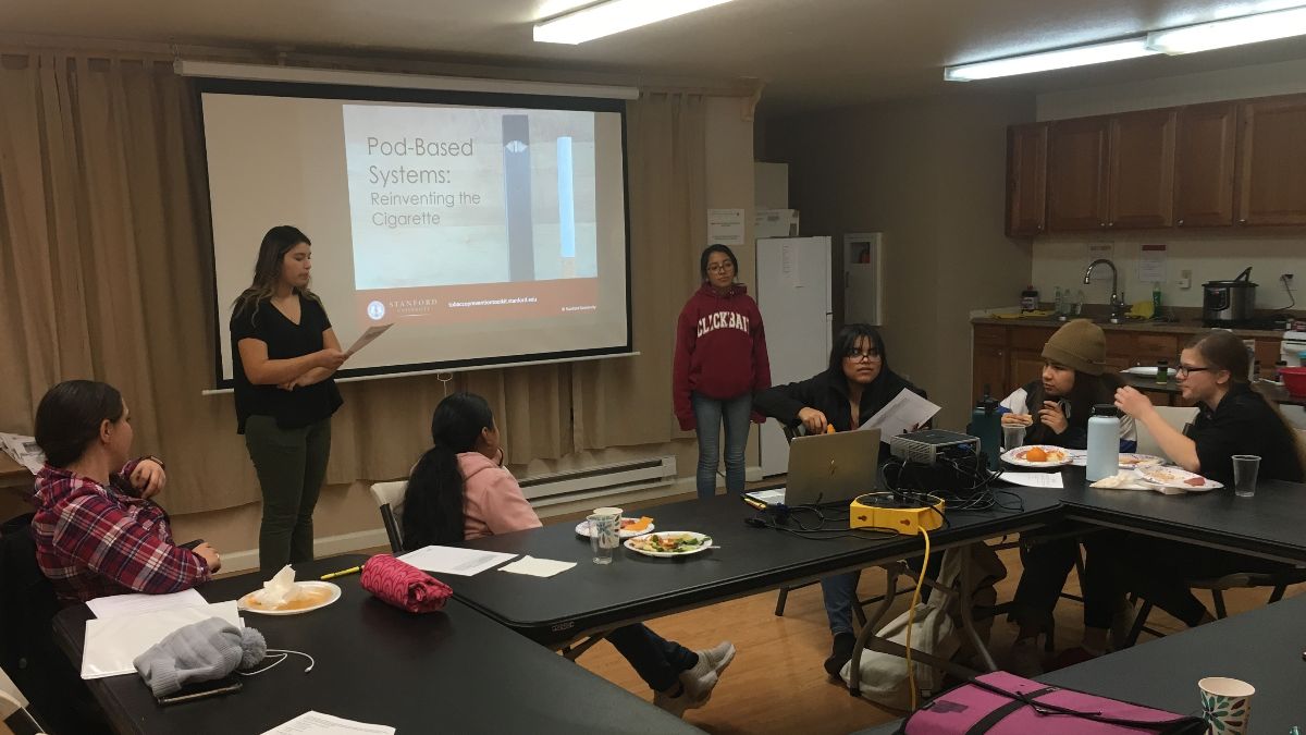 A young woman with a projected image consults notes in a room with seated youth sharing a meal.