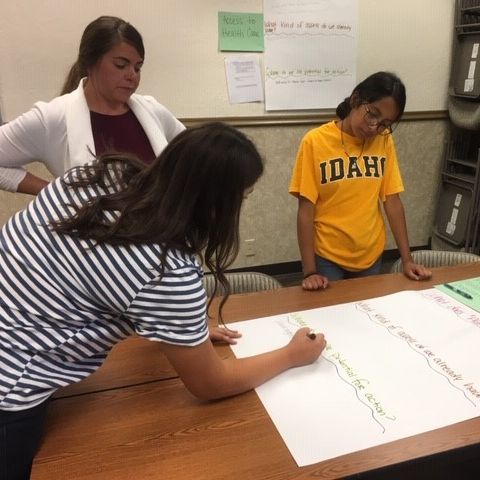 Three young women write with markers on a large paper.