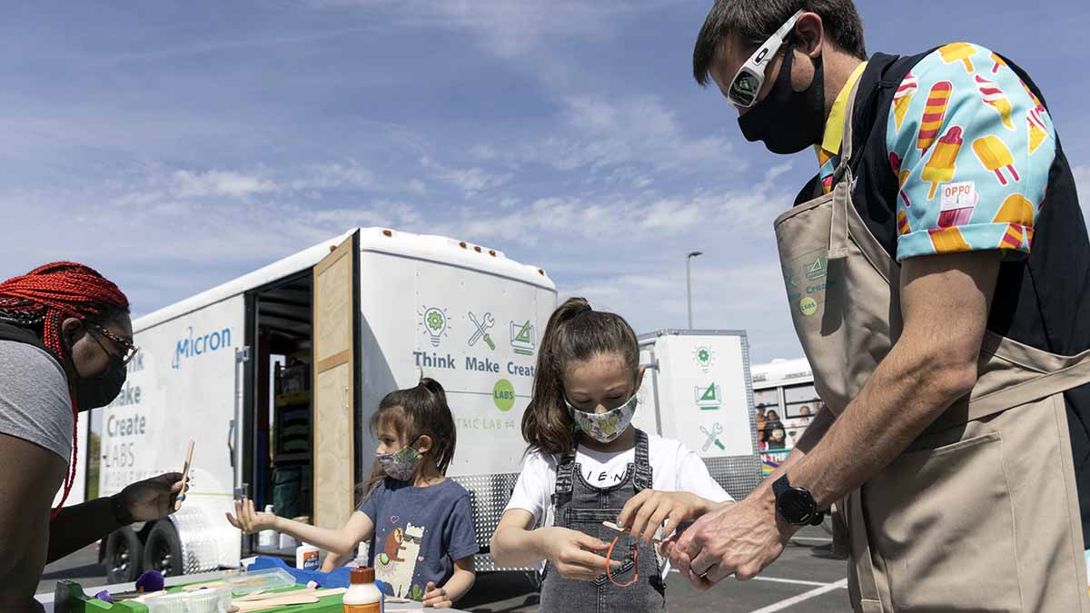 Two children work with popsicle sticks and rubber bands while two adults speak with them. They are outdoors near several trailers marked "Think, Make, Create Labs."
