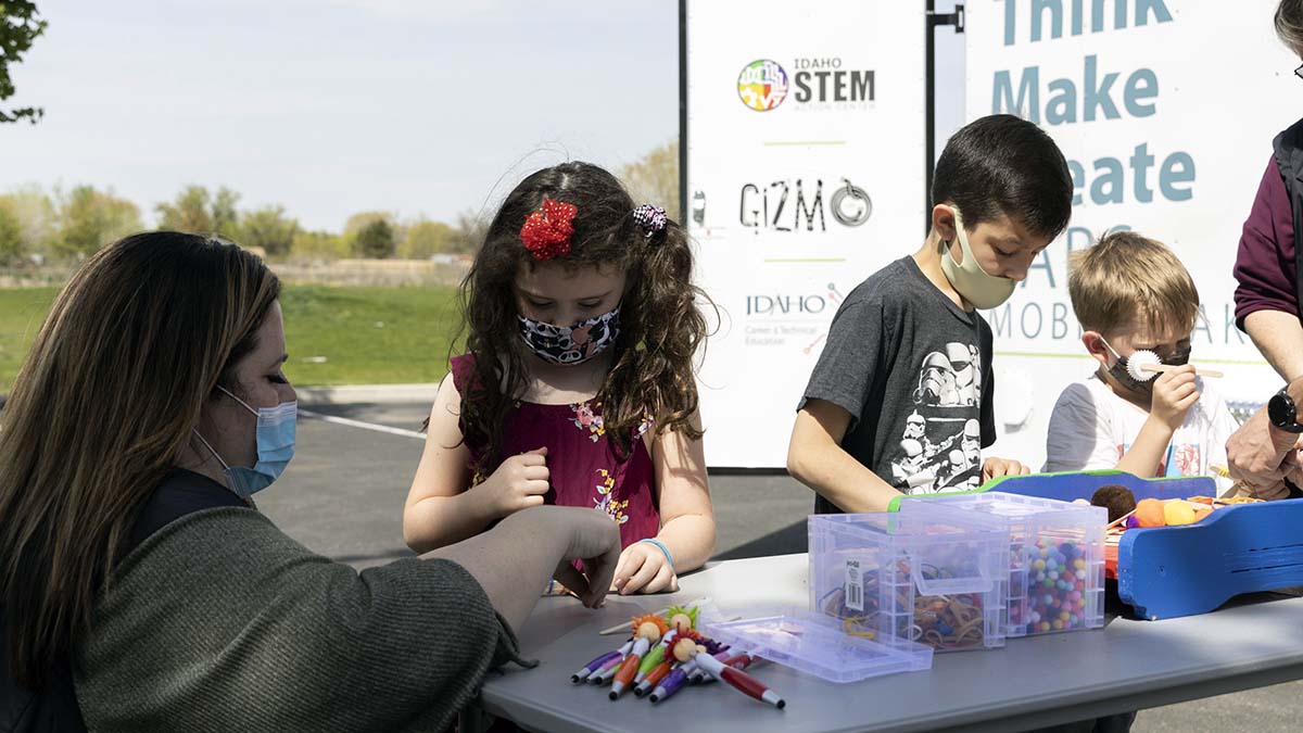 An adult holds a stick for a girl while another shows two boys crafts with pom poms.