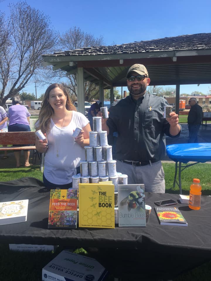 A woman and man hold up small containers outside. The table in front of them holds books about bees.
