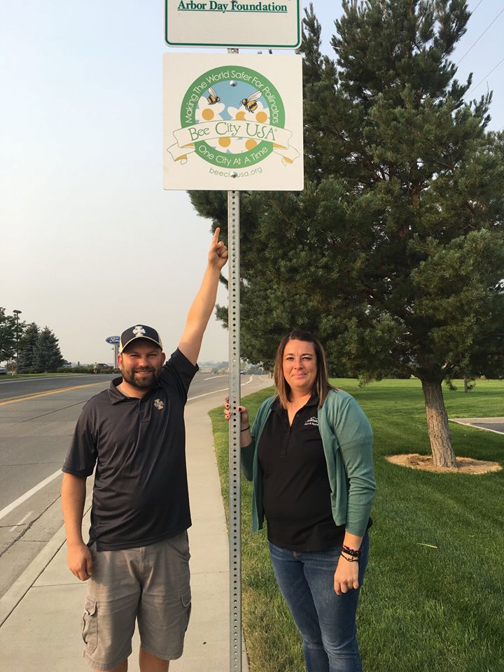 A man points to a street sign saying "Bee City USA" while a woman holds the signpole.