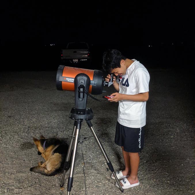 A young man peers into the eyepiece of a telescope at night.