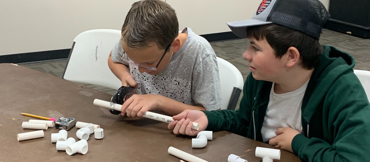 One boy cutting a PVC pipe with another boy holding the pipe