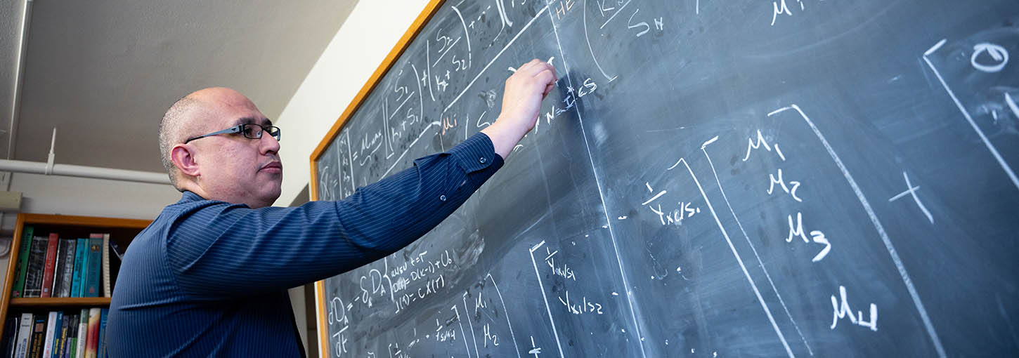 Man wearing glasses and blue shirt writes mathematical formula on blackboard.