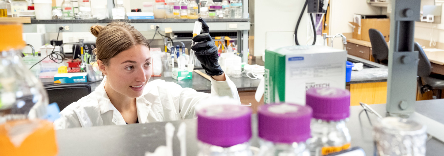 Woman peers at pipet through beakers in a science lab.