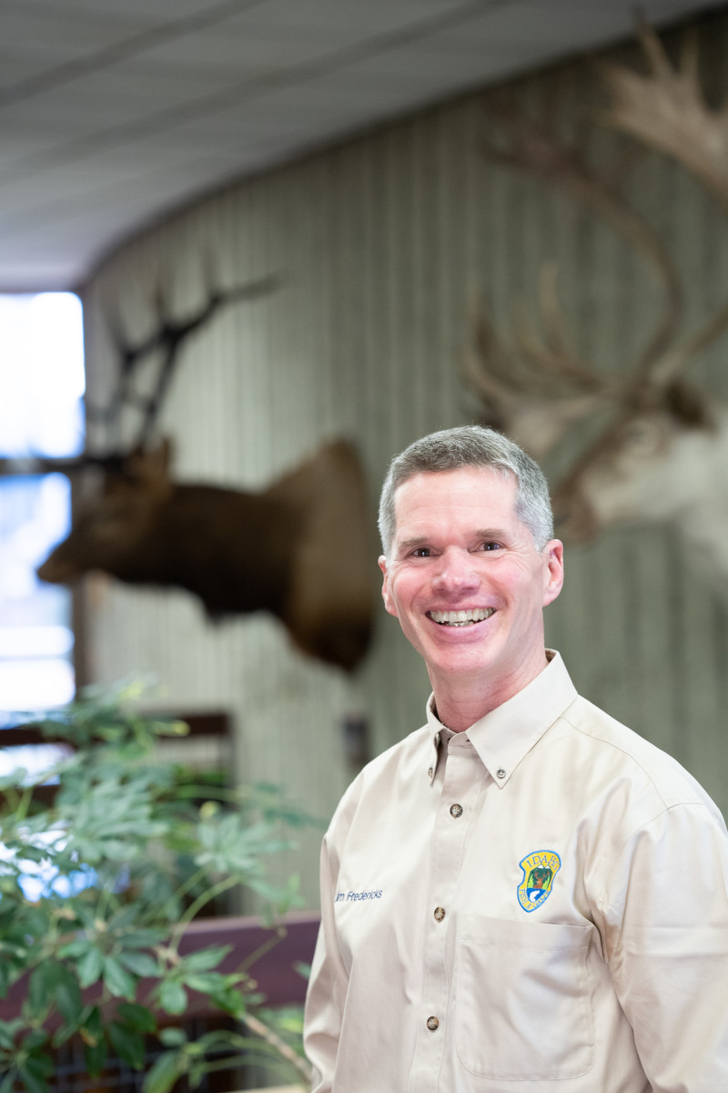 Smiling man stands in front of a mounted elk head.