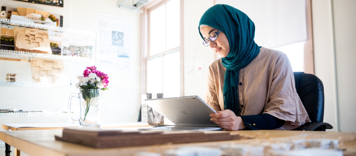 Layal Hussain works on her tablet at a wooden desk, surrounded by architectural design tools.