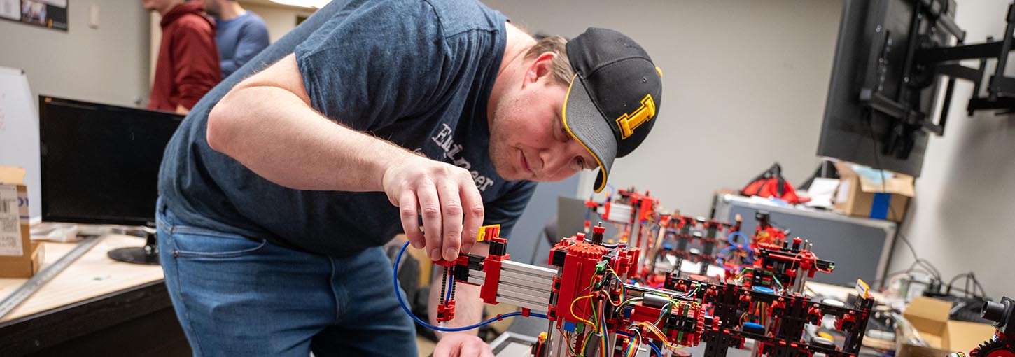 A young man in an Idaho baseball hat adjusts a piece on the arm of a robotic systems that simulates a mini factory in a robotics classroom.