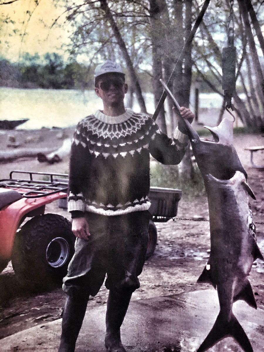 A man wearing a sweater displays a paddlefish near a river.