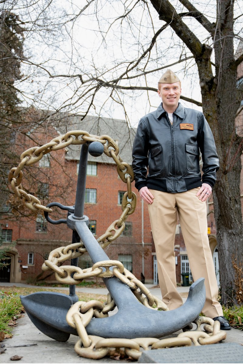 A naval officer in a leather jacket, beige slacks and a beige hat poses next to an anchor wrapped in a gold chain.