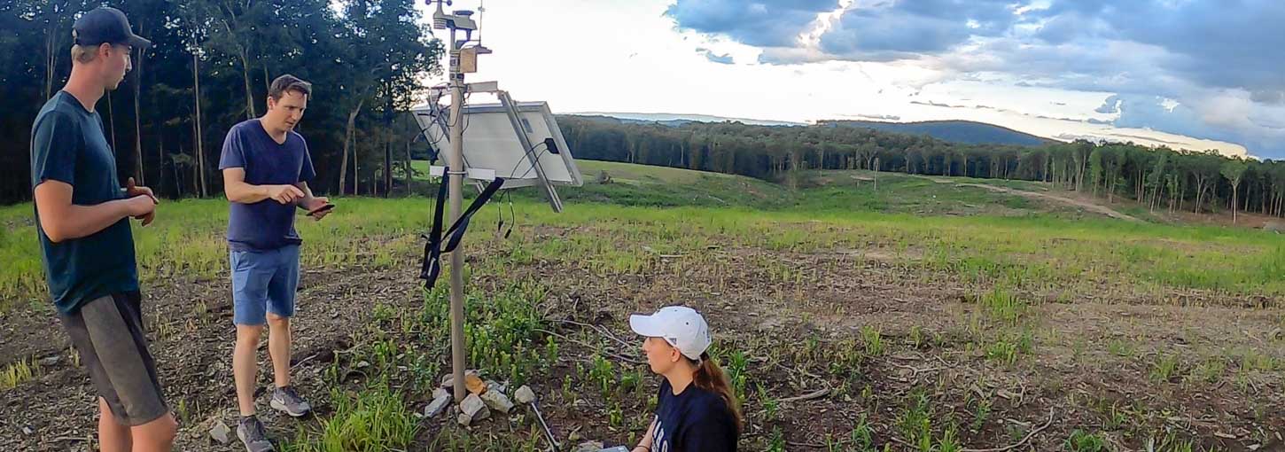 Three people stand on a hill in an open meadow near a miniature weather center surrounded by a forest.