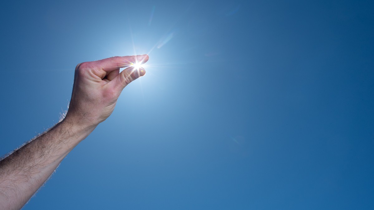 A person squints their fingers over the sun in a cloudless blue sky. 