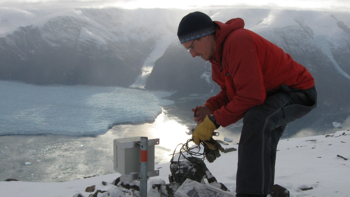 Tim Bartholomaus leans over a seismometer with a glacier and mountain behind him.