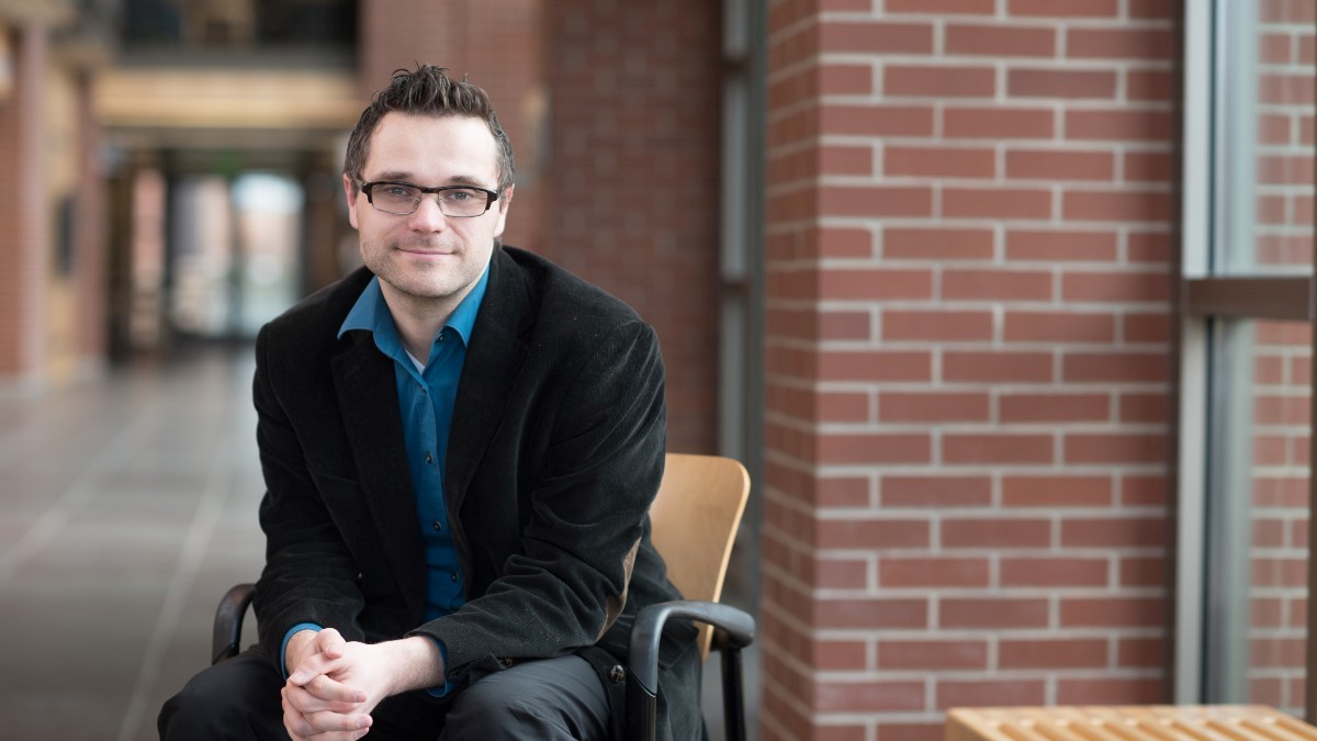 Professor Florian Justwan poses on a chair by a window down the hall from a long corridor during a sunny day.