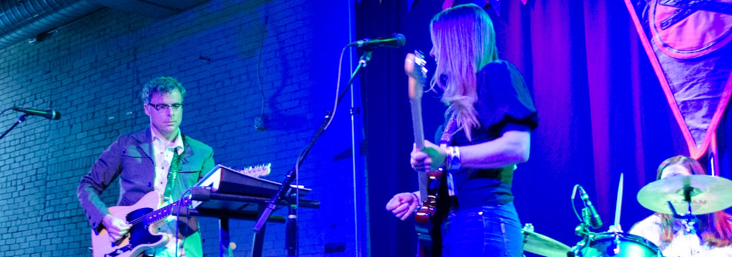 University of Idaho College of Law Professor Jason Dykstra, left, plays a beige electric guitar on a blue tinted stage, joined by another guitarist and drummer.