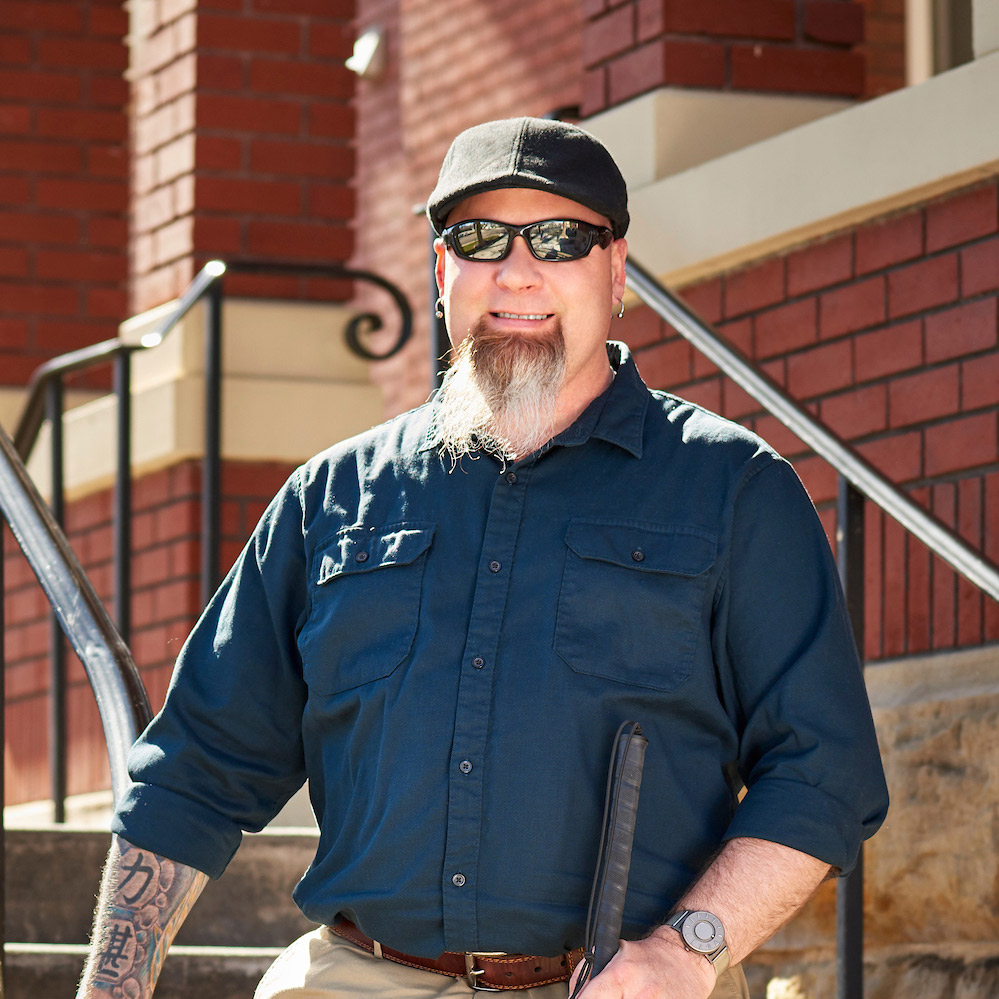 Earl Hoover stands on outdoor stairs, smiling at the camera on a bright day.