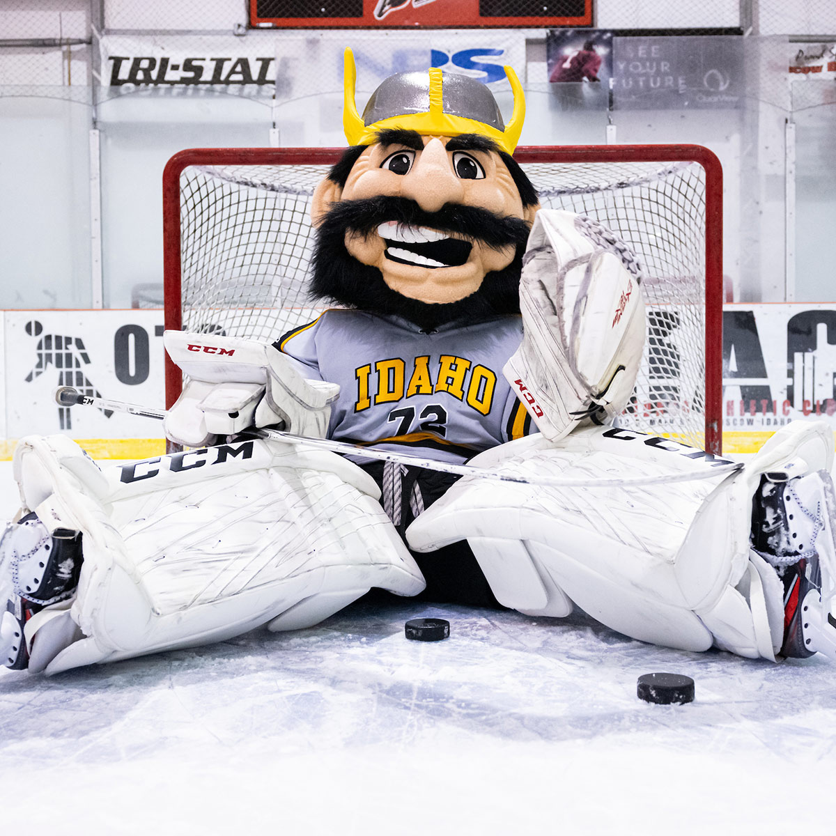 Joe Vandal sits defeated in front of the hockey goal, with pucks around him on the ice. He wears skates, goalie pads and gloves.
