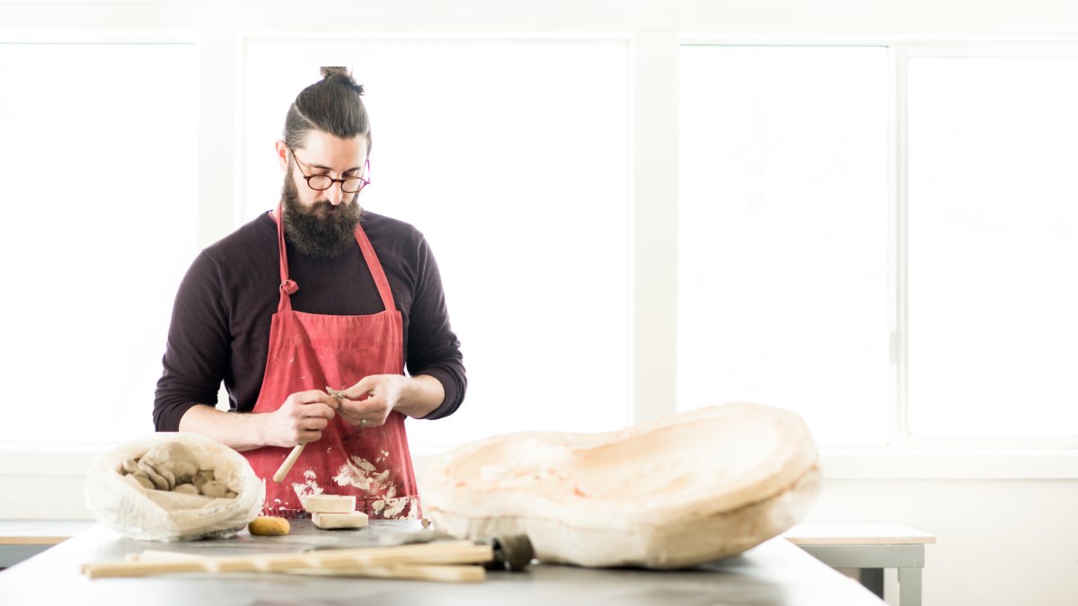 Casey Doyle stands at a table in a design studio working with a piece of clay.