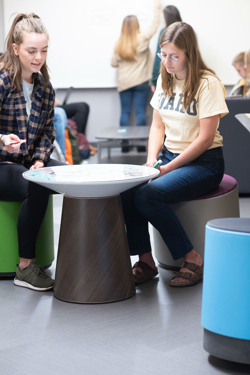 Two female student sit on colored stools at a whiteboard table.