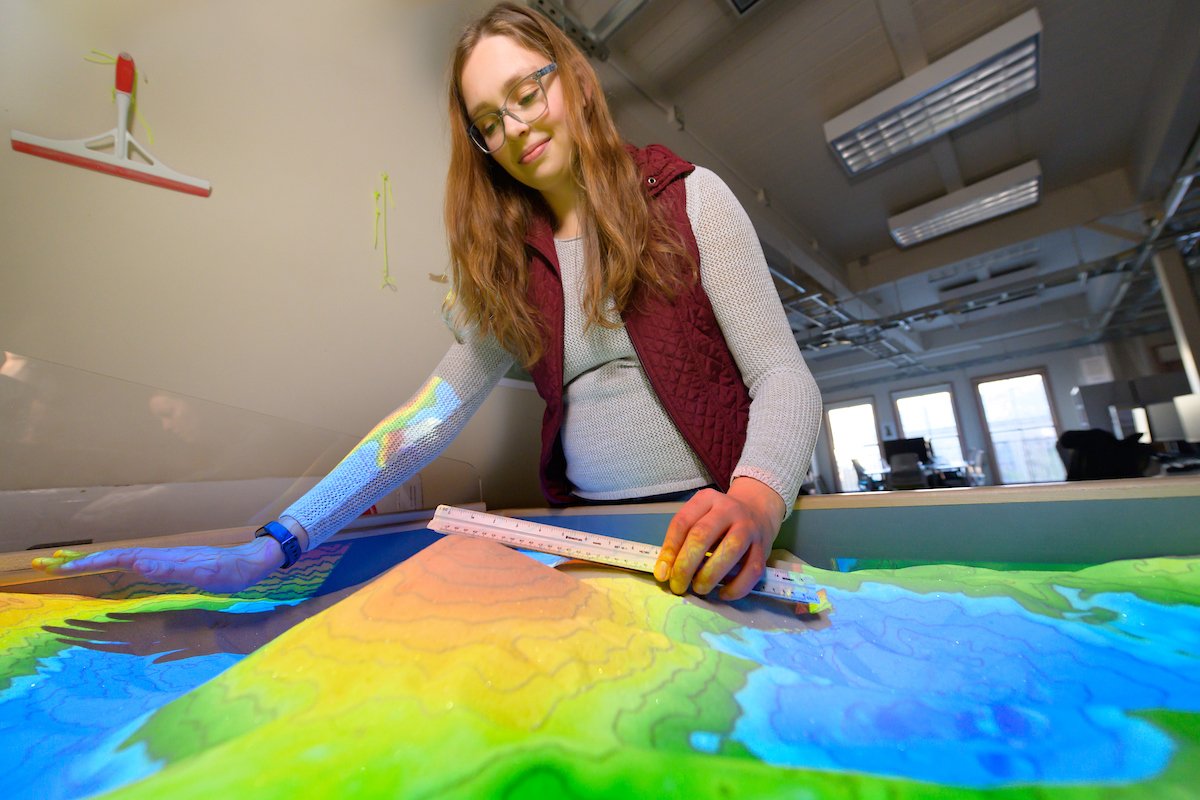 Katherine Woodhouse works at a table with edges that contains sand, with a map projected on it by colored light.
