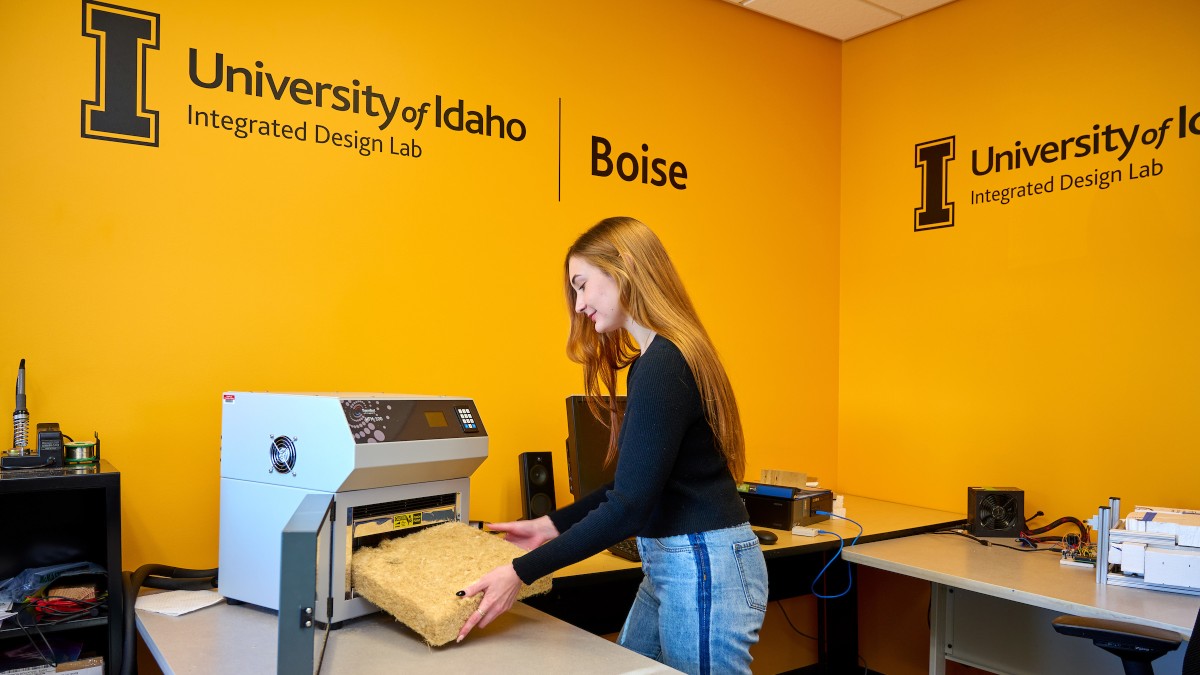 A female student uses equipment in the Integrated Design Lab. The wall behind her is pride gold stenciled with the logo of the University of Idaho Integrated Design Lab and the word Boise. 