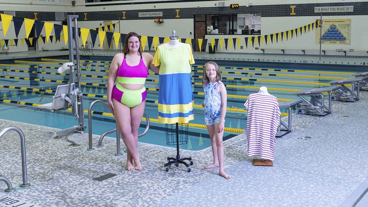 A woman and girl wearing swimsuits stand on the deck of a pool next to dress forms wearing swim coverups.