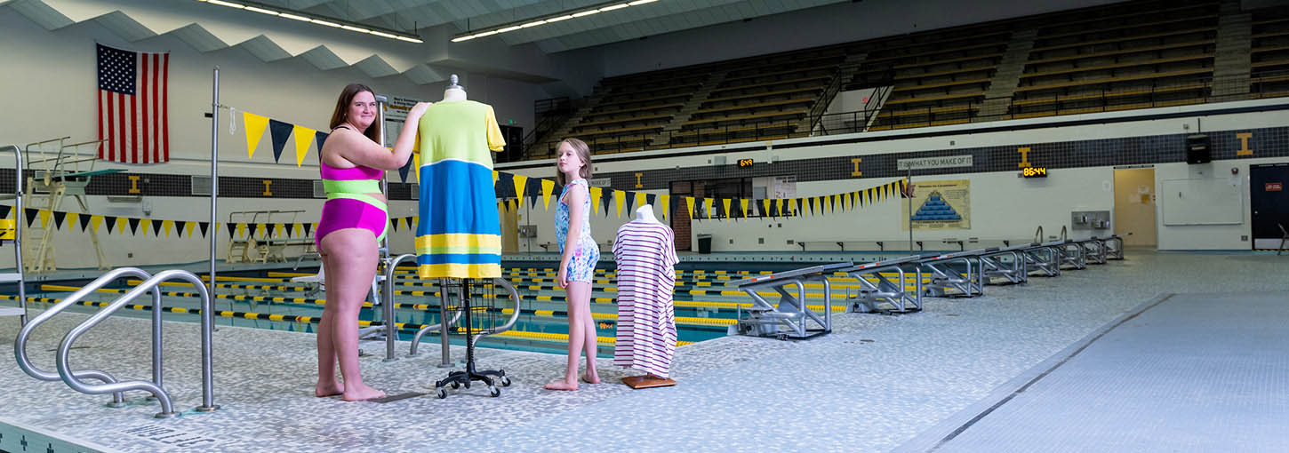 A woman and girl wearing swimsuits stand on the deck of a pool next to dress forms wearing swim coverups.