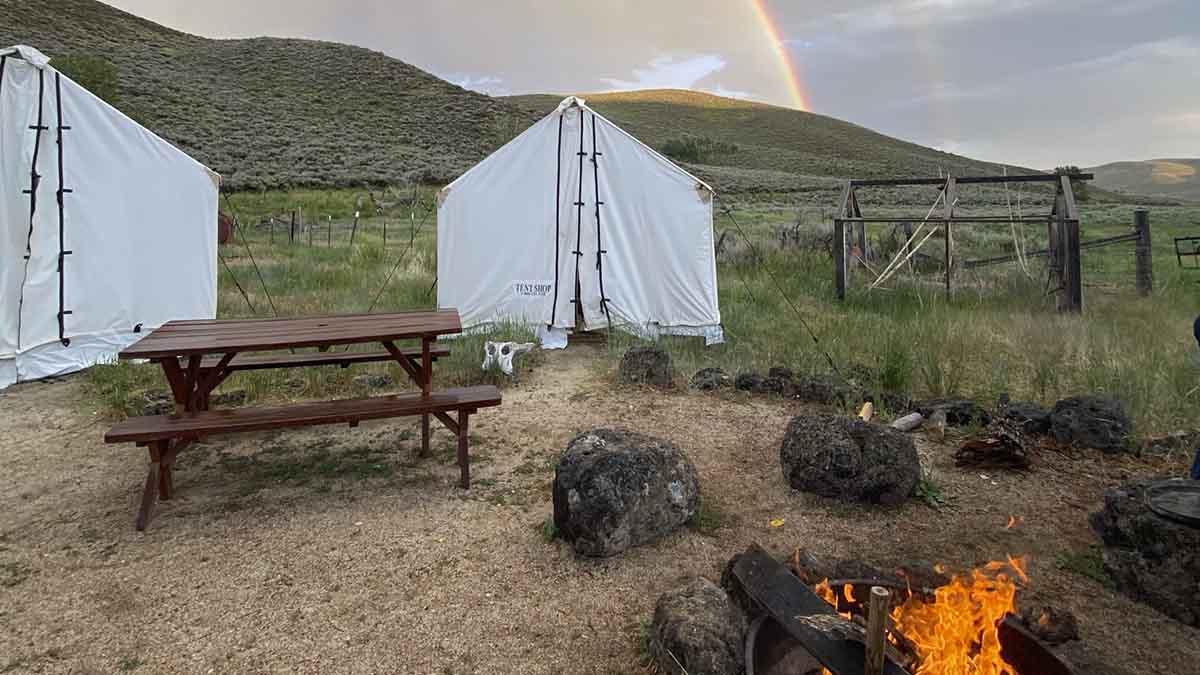 A white tent sits in front a picnic bench and firepit.