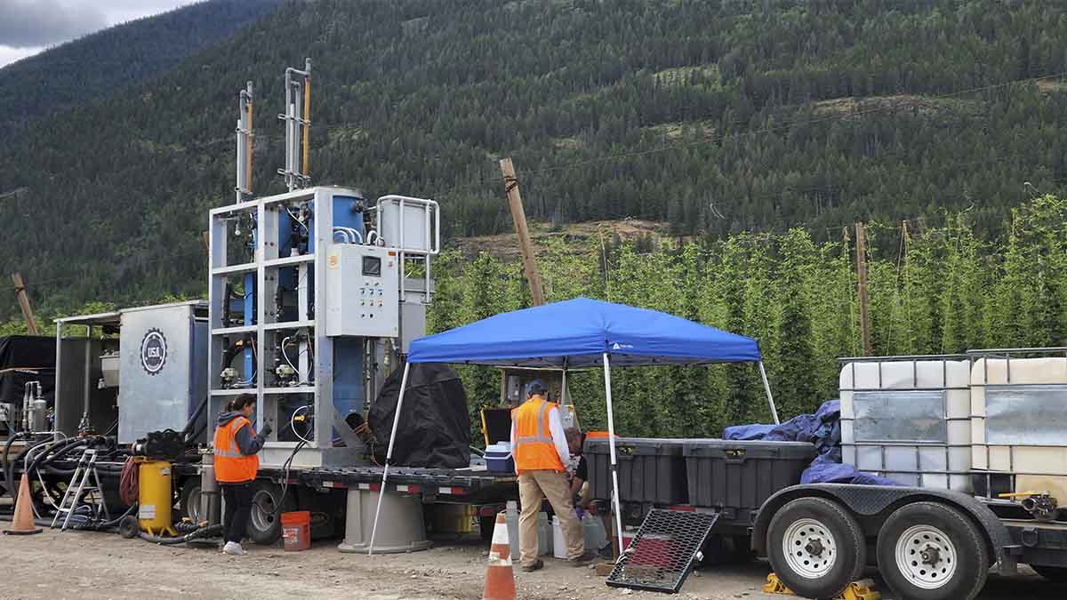 The Clean Water Machine sits on a trailer beside a row of hops while employees work.