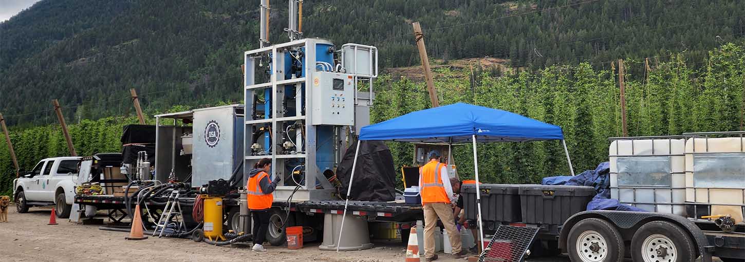 The Clean Water Machine sits on a trailer beside a row of hops while employees work.