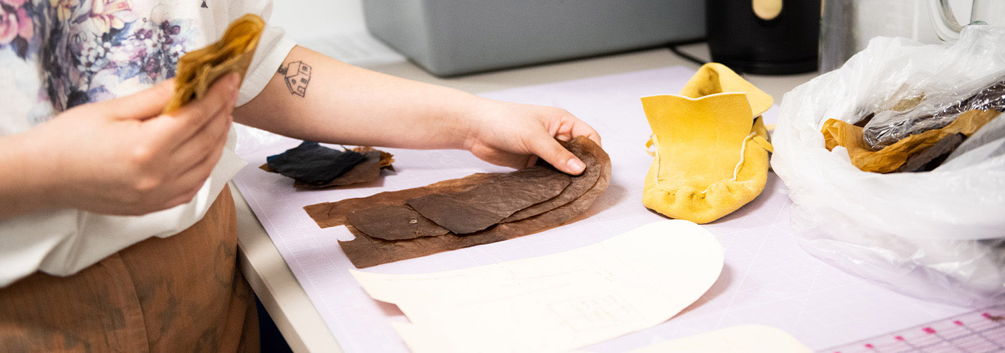 A person places cut out patterns of SCOBY leather on a table next to a pattern and leather moccasin.